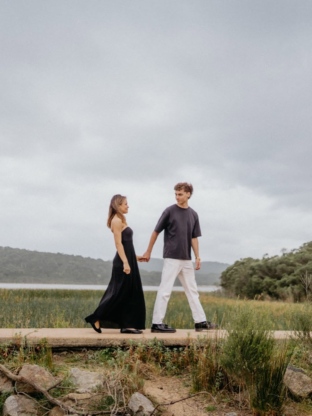 Sophie and Alex holding hands beside the lake beneath an open sky