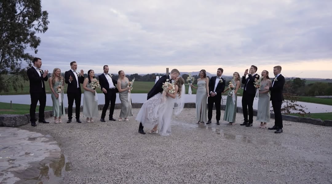 Wedding film still of a couple kissing, surrounded by their wedding party